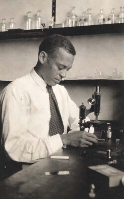 Black and white image of Dr. Ernest Everett Just in a laboratory setting. Just is seated at a desk with a microscope in front of him. His hands are adjusting a setting on the microscope that he is looking fown at. He is wearing a white dress shirt and tie and has his body facing at an angle to the camera.