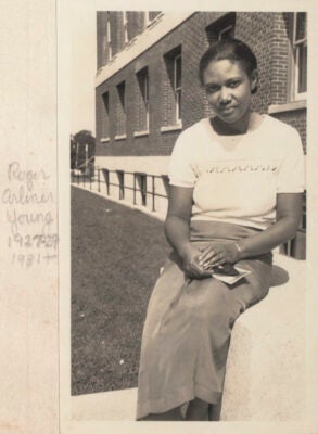 Black and white image of Dr. Roger Arliner Young seated on a low wall at the Woods Hole Oceanographic Institute campus. Young is looking at the camera, wearing a white shirt and long, dark skirt, and is holding a pamphlet in her hands crossed on her lap.