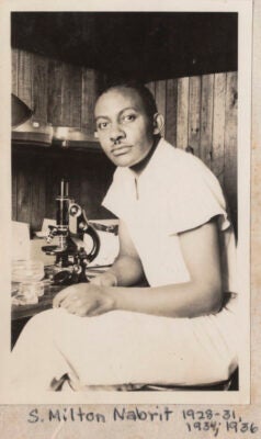 A black and white image of Dr. Samuel Milton Nabrit. Nabrit is seated at a small workstation with a microscope in front of him. He is looking at the camera, his body oriented to the left of the frame. There are glass dishes on the table to his left, and large glass dishes on a shelf above the table.